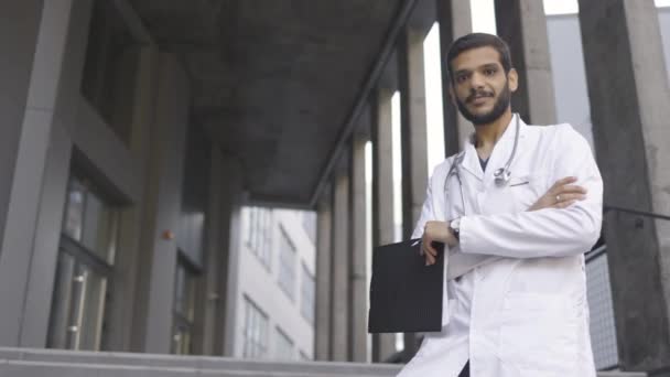 Lekarz w białej sukni, trzymający teczkę, stojący na schodach nowoczesnej kliniki na świeżym powietrzu, z kciukiem w górze. — Wideo stockowe