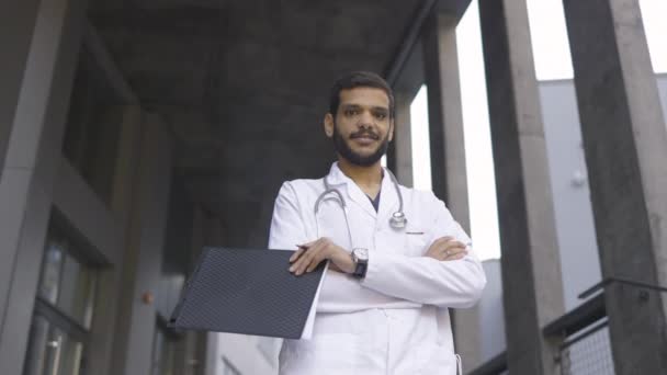 Médico árabe, segurando prancheta e caneta, de pé com os braços cruzados ao ar livre — Vídeo de Stock