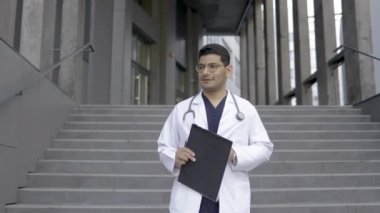 Arap erkek doktor, modern hastanenin merdivenlerinden iniyor, yana bakıyor ve notlar alıyor.