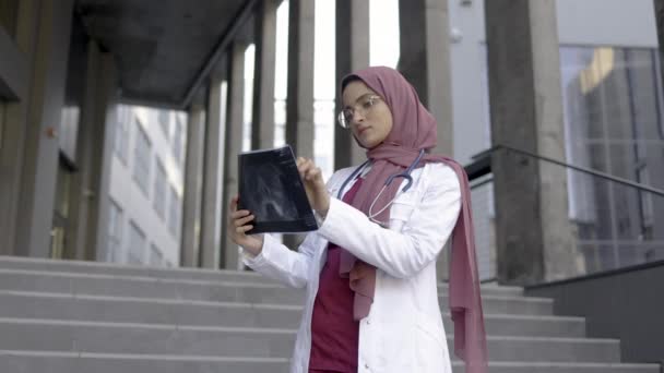 Médica muçulmana, de pé fora da clínica moderna nas escadas, e analisando a radiografia do crânio — Vídeo de Stock