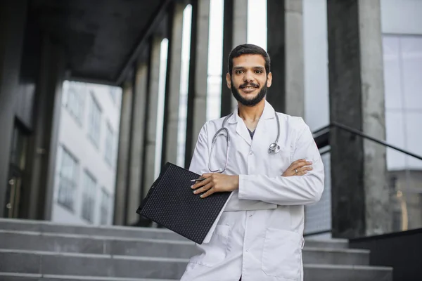 Arabische mannelijke arts, klembord en pen vasthoudend, buiten staande met armen gekruist — Stockfoto