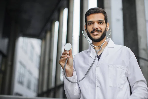 Sorridente studente di medicina araba maschile in camice bianco, in piedi fuori, mostrando il suo stetoscopio — Foto Stock