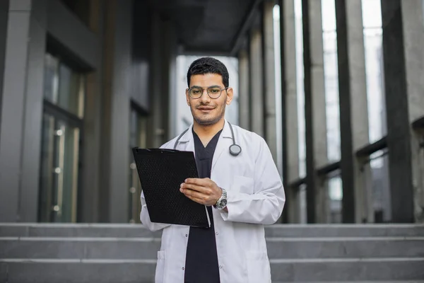 Arabo indiano medico maschile, in piedi fuori clinica moderna, prendendo appunti e riempiendo la cartella dei pazienti — Foto Stock