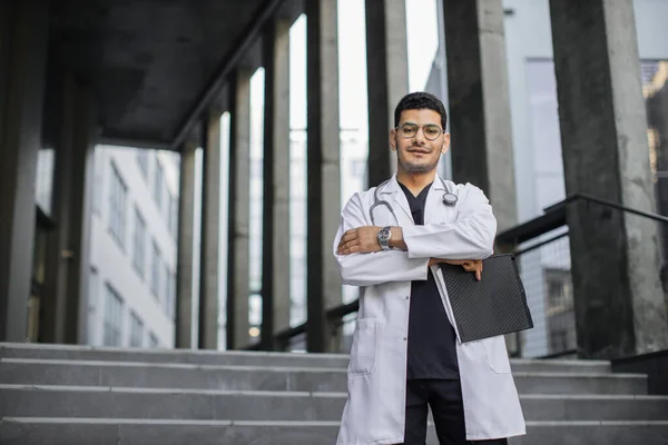 Arabian male physician, holding clipboard, standing with arms crossed outdoors clinic — Stock Photo, Image