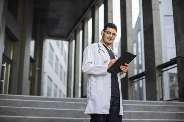 Arabo medico maschio, scendendo le scale fuori dal moderno ospedale, guardando la macchina fotografica e prendendo appunti — Foto Stock