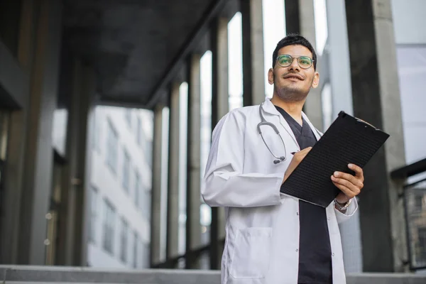 Professzionális férfi arab orvos írásban RX recept vágólapra, áll a kórház előtt — Stock Fotó