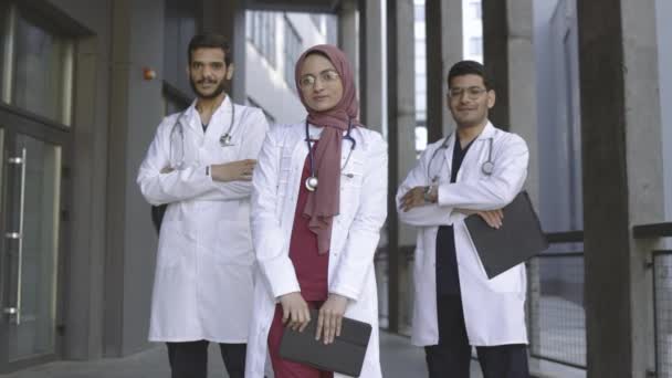 Tres jóvenes y confiados médicos árabes saudíes, de pie fuera del hospital y mirando a la cámara — Vídeo de stock