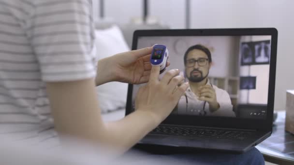 Жінка сидить перед ноутбуком і носить імпульсний оксиметр на свого пращура, розмовляючи з лікарем-чоловіком — стокове відео