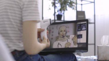 Gündelik giyinmiş bir kadın, Müslüman doktorun video blogunu izliyor, termometreyi nasıl kullanacağını anlatıyor.