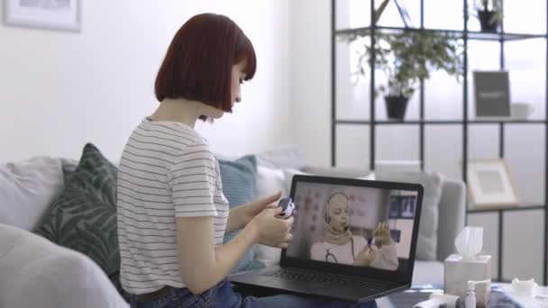 Vrouw met pulse oximeter in handen kijken onderwijs video van vrouwelijke arts op laptop — Stockvideo