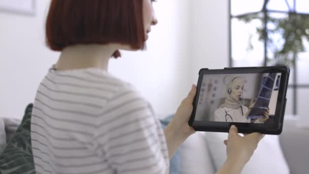 Жіночий пацієнт, сидячи вдома з планшетним ПК в руках, має відеочат зі своїм лікарем — стокове відео