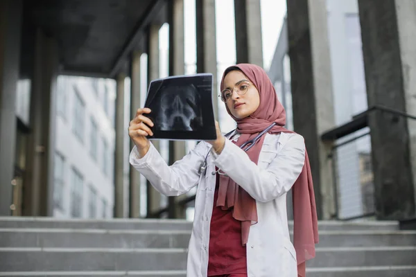 Muslimischer Arzt im Hijab, steht vor einem modernen Krankenhaus und betrachtet das Röntgenbild — Stockfoto