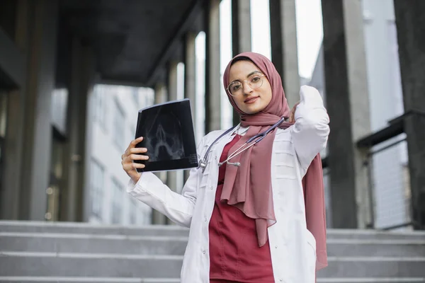 Young Arab woman physician in headscarf holding x-ray, standing outside modern clinic — Stock Photo, Image