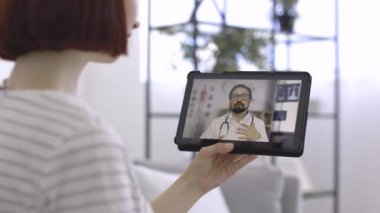 Kadın, dijital tablet kullanarak doktoruyla konuşurken el sallıyor.