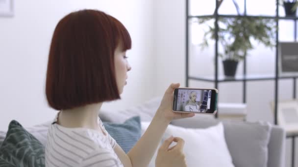 Femme, assise sur le canapé à la maison, et consulter son médecin en ligne à l'aide d'un smartphone — Video