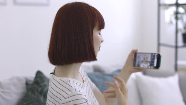 Jovem paciente, sentada no sofá em casa, tendo vídeo chat com médico árabe feminino no telefone — Vídeo de Stock