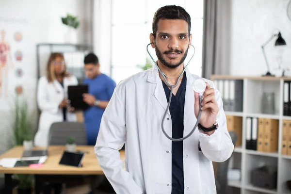 Medico di sesso maschile mediorientale in abito bianco, mostrando il suo stetoscopio alla telecamera — Foto Stock