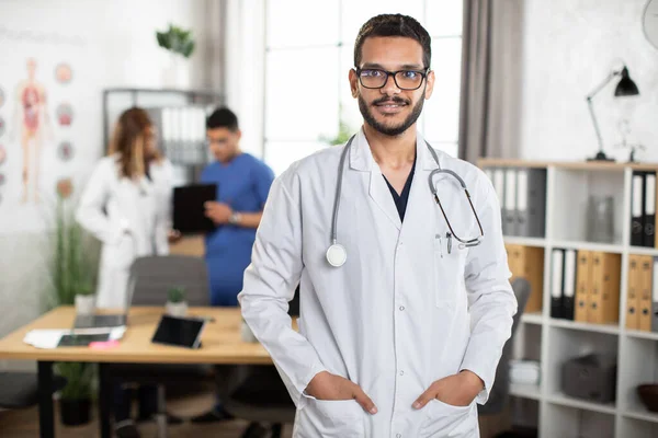 Medico arabo maschio con stetoscopio, che si tiene per mano nelle tasche del cappotto bianco, in piedi all'interno — Foto Stock