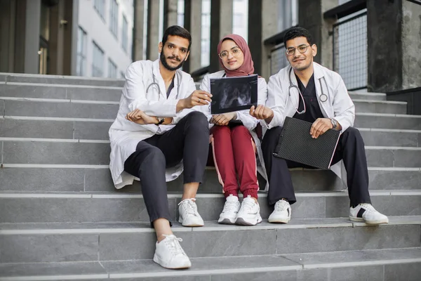 Drei arabische Arztpraktikanten sitzen draußen auf der Kliniktreppe und diskutieren über Schädel-Röntgen — Stockfoto