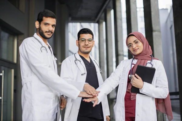 Two male handsome doctors and their pretty female colleague in hijab, stacking hands — Stock Photo, Image