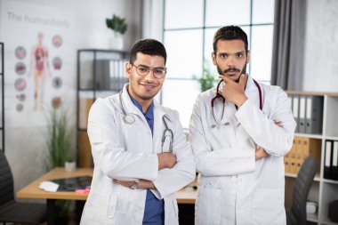 İki Arap doktor, klinikte kollarını kavuşturup kameraya bakıp gülümsüyor..