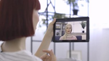 Başörtülü Müslüman kadınla tablet bilgisayar ekranı, çevrimiçi görüntülü sohbet yoluyla Kafkasyalı arkadaşıyla konuşuyor