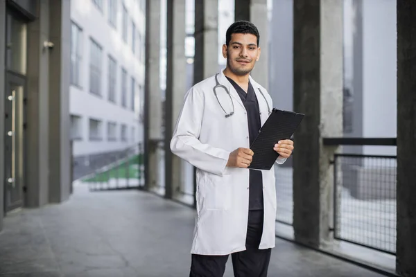 Medico maschio indiano in camice bianco, in piedi con appunti in mano all'aperto — Foto Stock