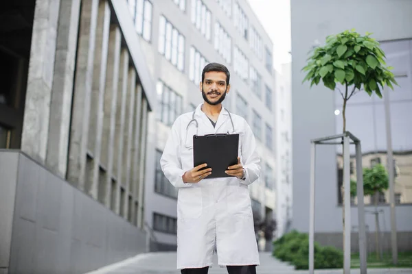 Operatore sanitario arabo in cappotto bianco, in piedi con cartellina appunti in mano all'aperto — Foto Stock