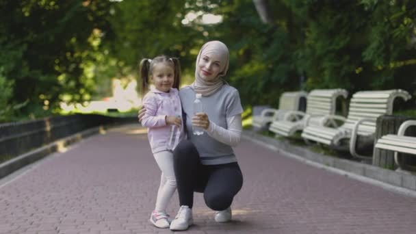 Muslim mother in hijab, posing with her cute daughter holding bottles with water, outdoors in park — Stock Video