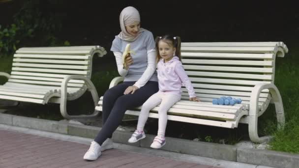 Мусульманская мать кормит свою маленькую милую дочь бананом, сидя вместе на скамейке в парке — стоковое видео