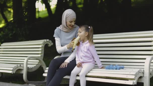 Arab muslim mom and her cute little daughter, eating banana resting on a bench in the public park. — Stock Video