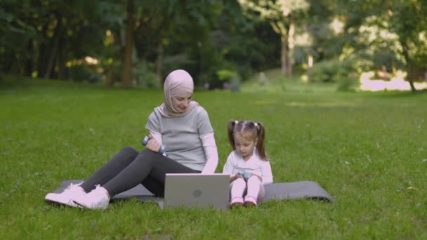 Maman en hijab montrant à son enfant comment faire de l'exercice avec des haltères, regarder la vidéo sur ordinateur portable dans le parc — Video