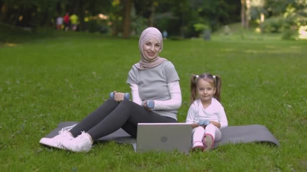 Arabska matka w hidżabie i 4-letnia córka, siedząca na wycieraczce w parku i podnosząca hantle — Wideo stockowe