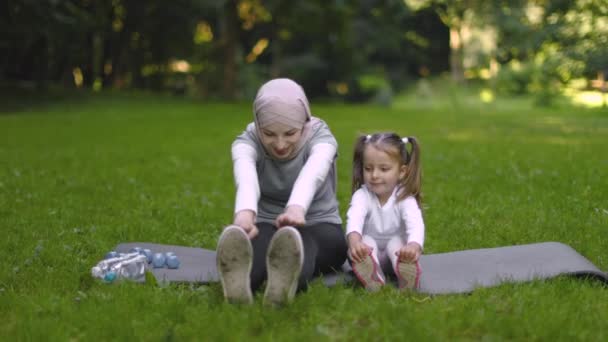 Liten flicka och hennes sportig Muselman mamma är gör stretching övningar i park sitter på grå matta — Stockvideo