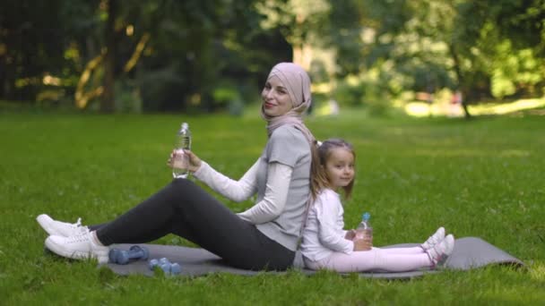 히잡에 살고 있는 회교도 여자, 공원에서 딸 과 함께 매트 위에 앉아 물을 마시는 모습 — 비디오