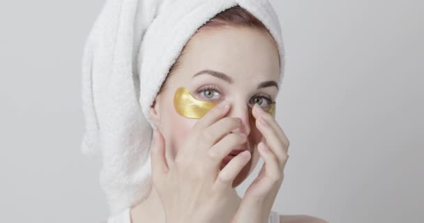 Nádherná usmívající se žena s ručníkem na hlavě nanášení zlaté oční pásky během domácí lázeňské procedury — Stock video