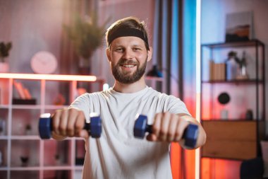 Evde antreman yapmak için halterleri kullanan kafa bantlı gülen adam.