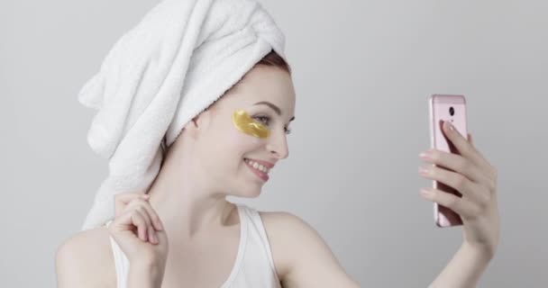 Lady with golden eye patches and towel on head, making selfie on cellphone or having video call — Stock Video