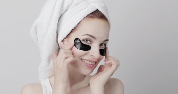 Lady with perfect clear skin, and hair wrapped in towel, applying black collagen under eye patches — Stock Video