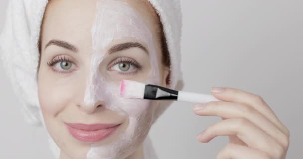 Mujer con toalla en la cabeza aplicando mascarilla facial en la cara, usando cepillo cosmético, sobre fondo gris. — Vídeo de stock