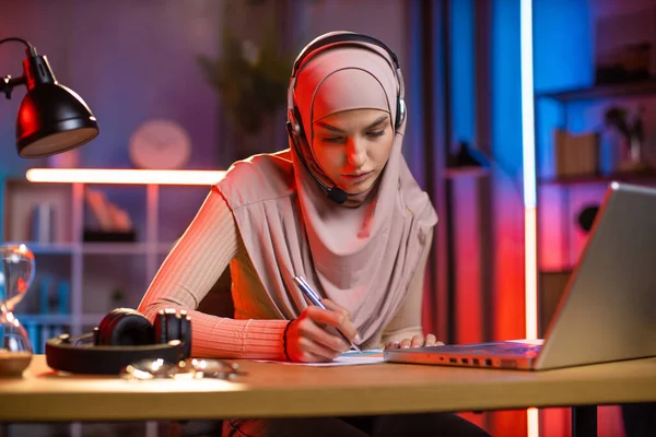 머리 스카프와 헤드셋을 하고 있는 아라비아 여성, 노트북에서 온라인 웹툰을 들으면서 필기를 하고 있는 모습 — 스톡 사진