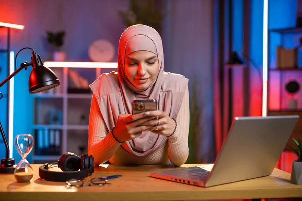 히잡에 앉아 저녁에 집에서 다채 로운 조명을 받는 방에 앉아 스마트폰을 사용하는 이슬람 여성 — 스톡 사진