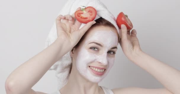 Mulher bonita com toalha na cabeça e máscara facial, se divertindo, sorrindo e segurando dois pedaços de tomate — Vídeo de Stock