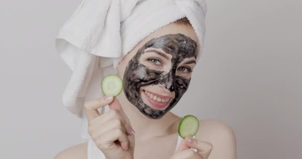 Menina feliz com toalha na cabeça, e máscara facial preta, posando com duas peças de pepino nas mãos — Vídeo de Stock