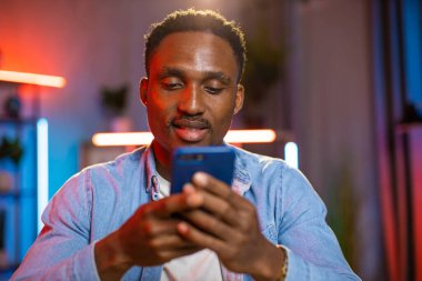 Siyah tenli bir adam akşamları evde dinleniyor ve akıllı telefondan arkadaşlarıyla sohbet ediyor.