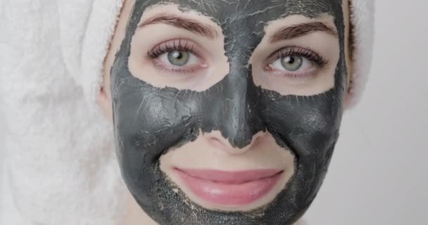 Mujer joven con la máscara facial de barro de barro negro seco en la piel, mirando a la cámara, sonriendo y guiñando el ojo — Vídeos de Stock