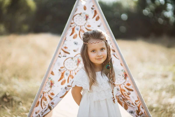Menina muito pequena, vestindo elegante vestido branco e penas acessórios de cabelo de pé perto teepee — Fotografia de Stock