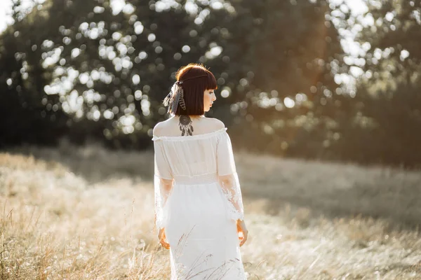 Mulher indígena americana, de vestido branco, tatuagens e cabelo curto com penas, andando em campo — Fotografia de Stock