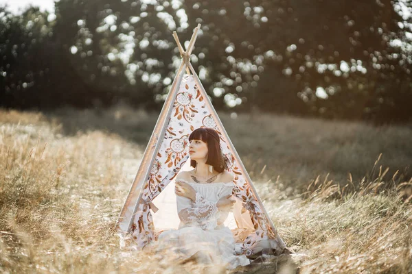 Hippie mulher vestindo boho vestido e penas acessórios de cabelo, sentado perto da tenda wigwam — Fotografia de Stock
