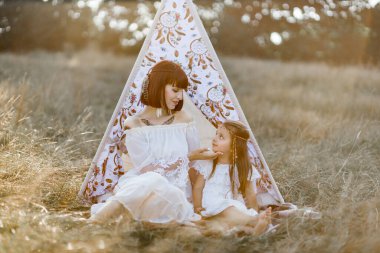 Anne küçük kızının yüzüne hafifçe dokunur, yaz tarlasında çadırın yanında birlikte oturur.